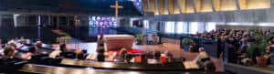 Panoramic view of a church interior with a modern design. Congregants sit in pews facing an altar. A large cross is displayed at the center, surrounded by colorful stained glass windows. The walls are concrete with a geometric pattern and potted plants decorate the space.