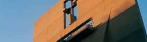 A modern, angular building facade is bathed in warm light. The structure features geometric cutouts and a balcony with decorative elements casting shadows on the surface. The sky is clear and blue, enhancing the warmth of the light on the building.