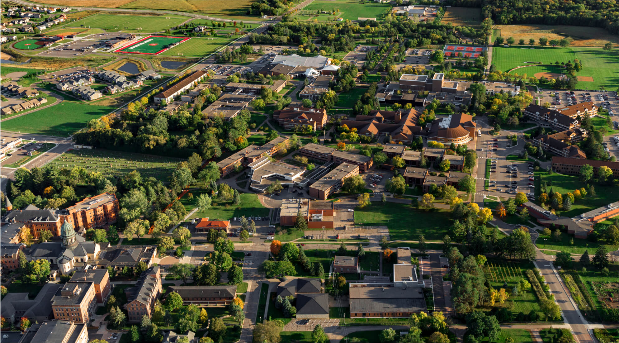 Aerial view of a sprawling college campus with numerous buildings, parking lots, sports fields, and green spaces. Trees and greenery surround the area, and a mix of academic, residential, and recreational buildings are dispersed throughout the landscape.