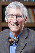 A smiling elderly man with short white hair and glasses is wearing a gray checkered suit jacket, a blue shirt, and a patterned bow tie. He stands in front of shelves filled with colorful books.