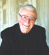 An elderly man with short white hair wearing glasses is smiling. He is dressed in a dark suit jacket and appears to be in front of a light-colored wall with a textured surface.