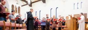 A group of people stands in a well-lit church with arched windows, holding red hymnals and singing. A priest stands at a podium on the right, and a religious figure in black robes stands among the congregation. The setting suggests a service or choir practice.