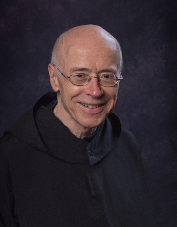 An elderly man with a bald head and glasses, dressed in a black robe, smiles warmly against a dark, textured background.