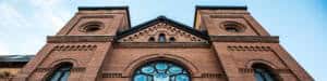 A low-angle view of a large, historic brick building featuring architectural details such as circular and arched windows, intricate brickwork, and twin towers under a clear blue sky.