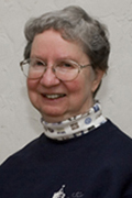 An elderly person with short gray hair is smiling. They are wearing glasses, a dark-colored sweater, and a light-colored turtleneck with a pattern. The background is a plain, light-colored wall.