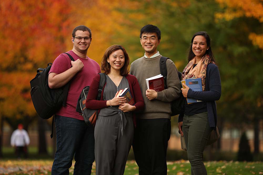Four smiling people standing outdoors in front of autumn trees, holding books and wearing casual clothes. They appear to be students, with backpacks on their shoulders. The background features vibrant fall foliage in shades of red, orange, and green.
