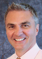A man with short gray hair wearing a light pink shirt and a light brown tie is smiling at the camera. The background is a mottled blue and gray backdrop.