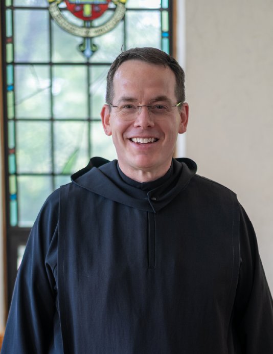 A man wearing glasses and a black monastic robe smiles in front of a stained glass window depicting various colors and patterns.