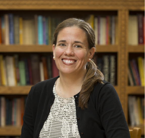 A woman with long, light brown hair tied back in a ponytail is smiling. She is wearing a black cardigan over a patterned blouse and has a necklace. Behind her is a wooden bookshelf filled with a variety of books.