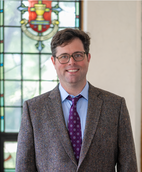 A man wearing glasses, a brown textured suit jacket, a light blue shirt, and a purple patterned tie is standing and smiling. A colorful stained glass window featuring a shield emblem is in the background.