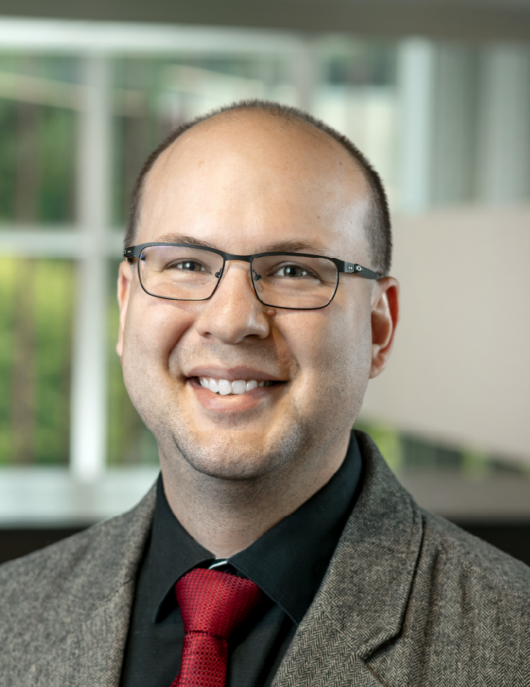 A man with a bald head and glasses is wearing a gray blazer, a black shirt, and a red tie. He is smiling and standing indoors with a blurred background featuring large windows.
