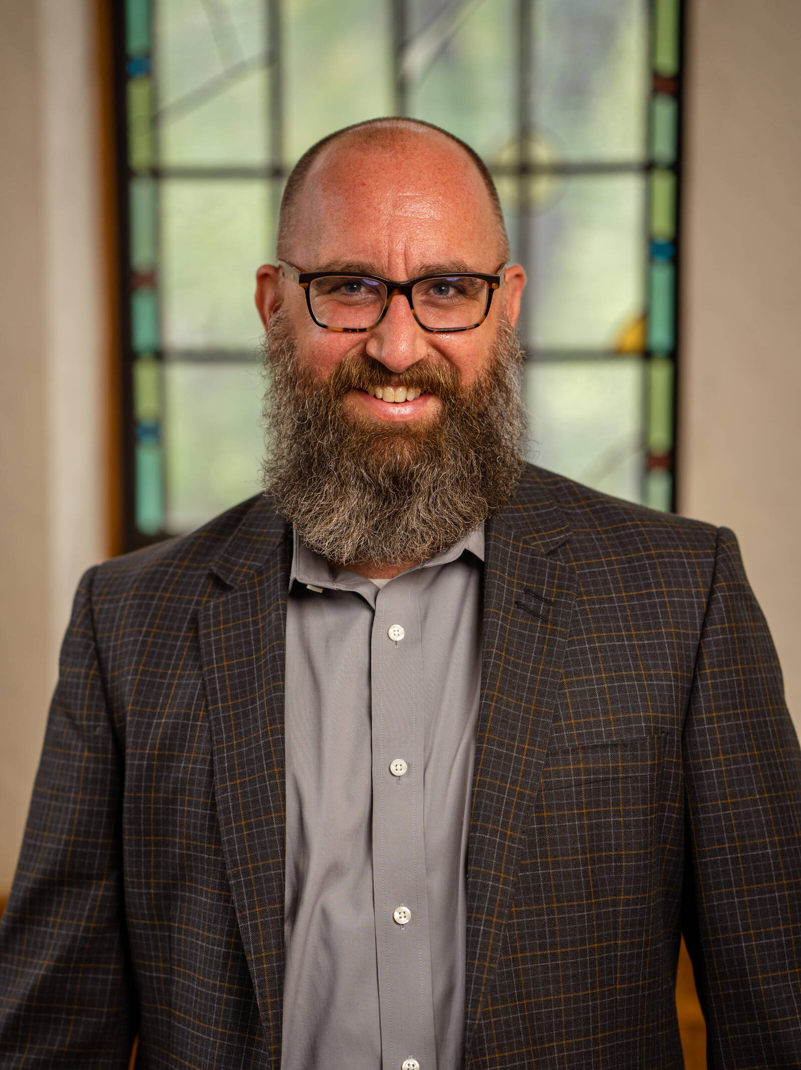 A smiling man with glasses, a bald head, and a full gray beard wears a plaid suit jacket and gray shirt, standing indoors in front of a blurred stained glass window.