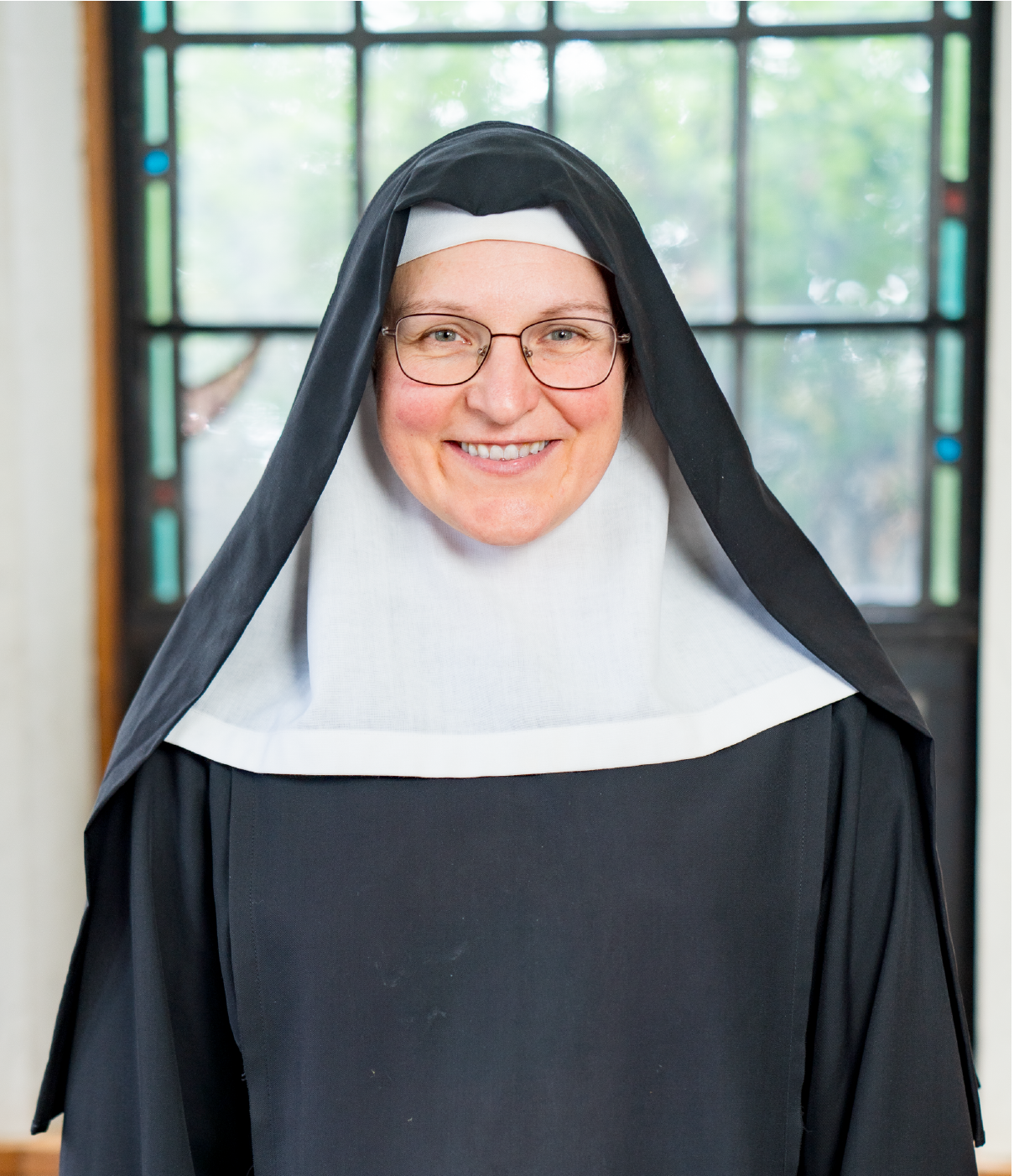 A smiling woman dressed in a traditional black and white nun's habit, including a veil and wimple, stands in front of a window with colored glass. She also wears glasses and is standing indoors with soft natural light illuminating her face.