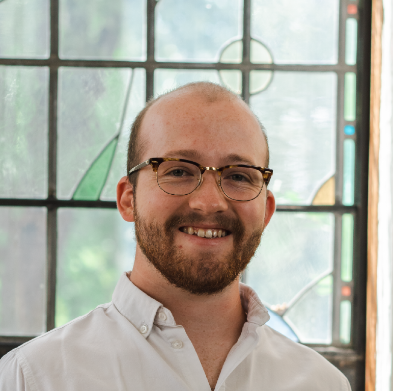 A man with short hair, a beard, and glasses is smiling while wearing a white button-up shirt. He is standing in front of a window with stained glass panels.
