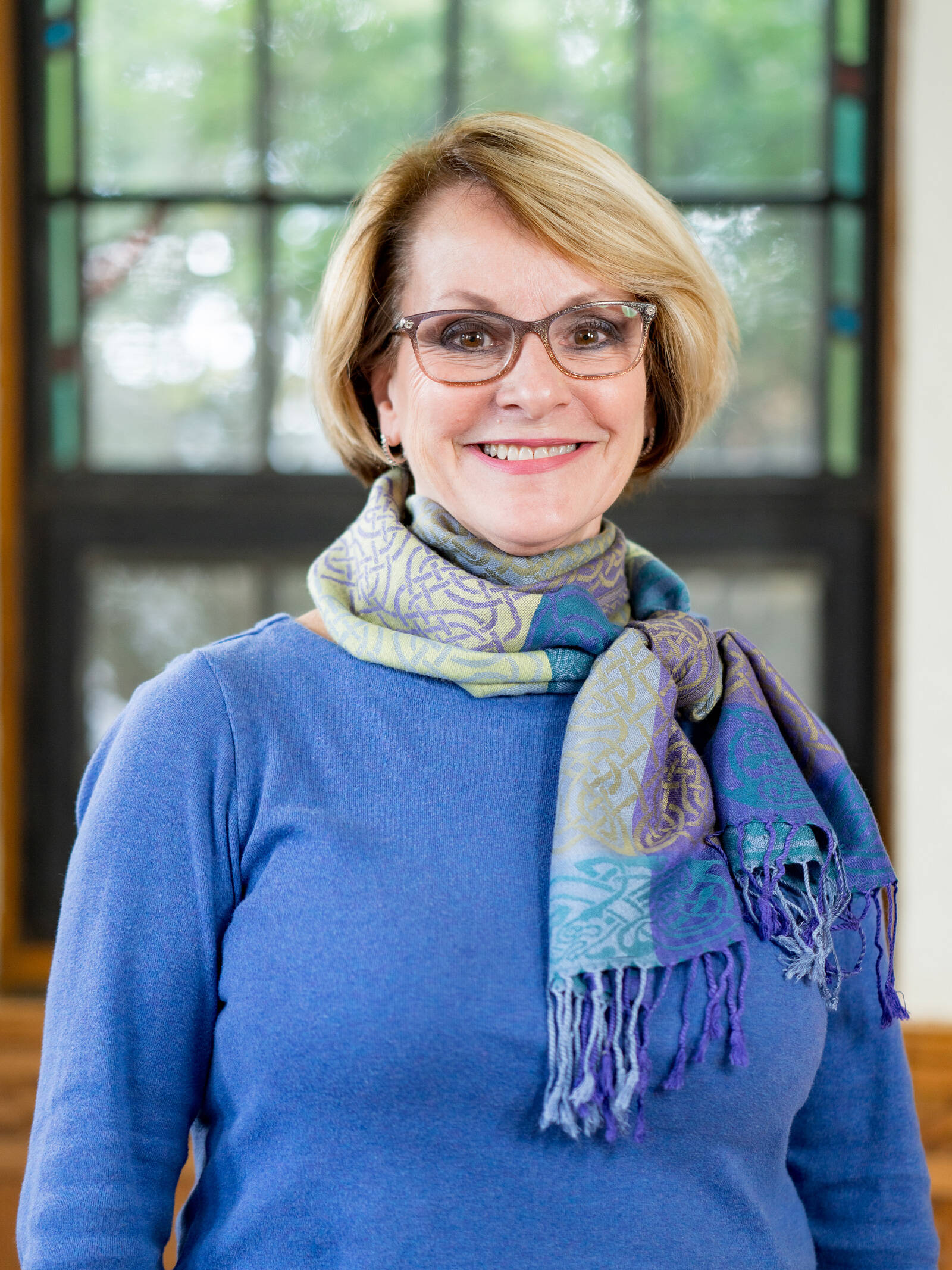 A smiling older woman with short blonde hair and glasses, wearing a blue sweater and a multicolored scarf, stands indoors in front of a window with decorative panes.