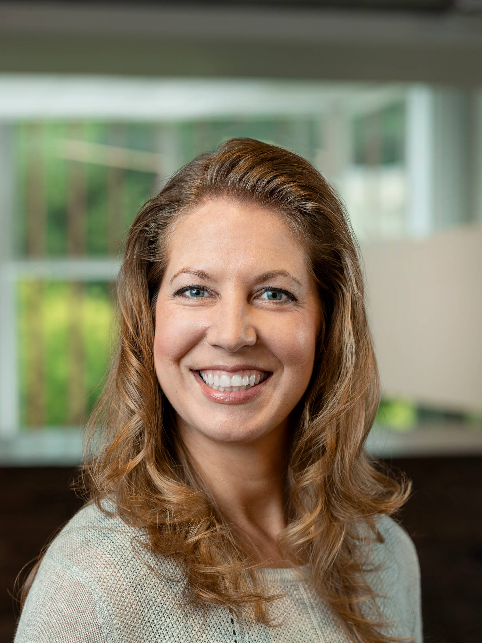 A woman with wavy, light brown hair and a light sweater smiles at the camera in a well-lit indoor setting with a blurred background of windows and greenery.