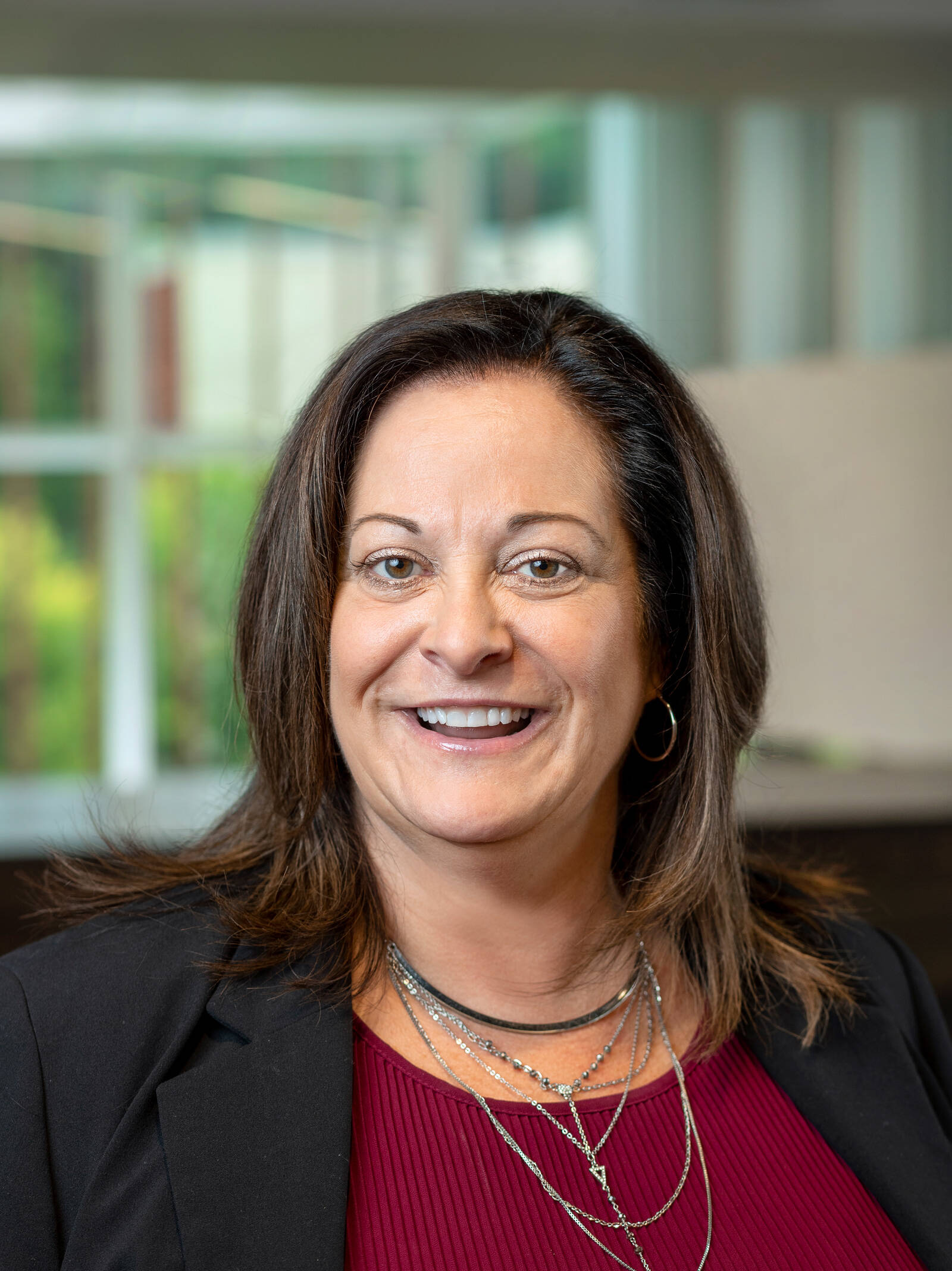 A woman with shoulder-length brown hair, wearing a black blazer over a maroon top and layered silver necklaces, smiles while standing indoors with large windows in the background.
