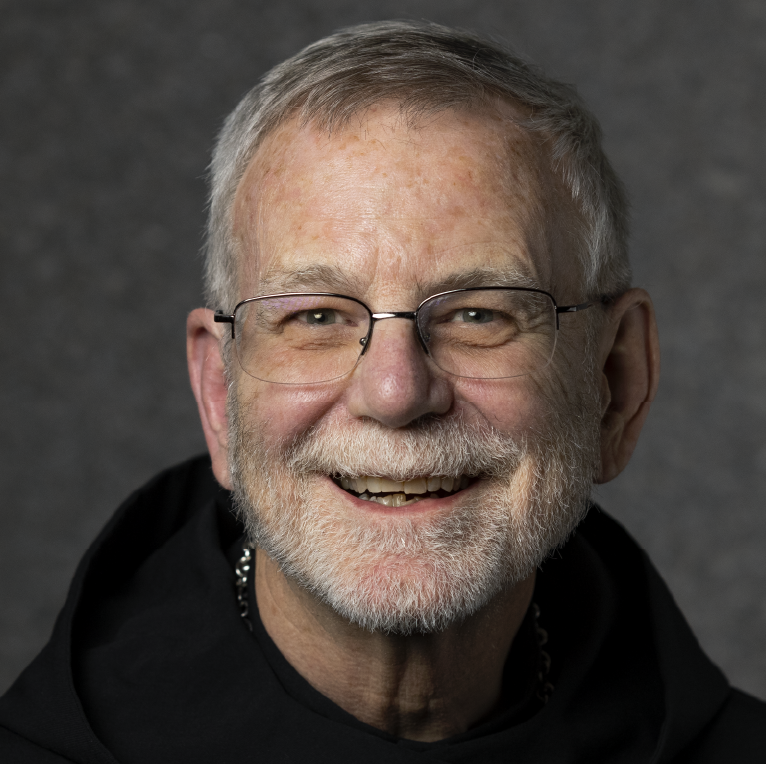 An elderly man with glasses, a white beard, and short gray hair smiles warmly. He is wearing a black hooded garment and a chain necklace. The background is a textured gray.