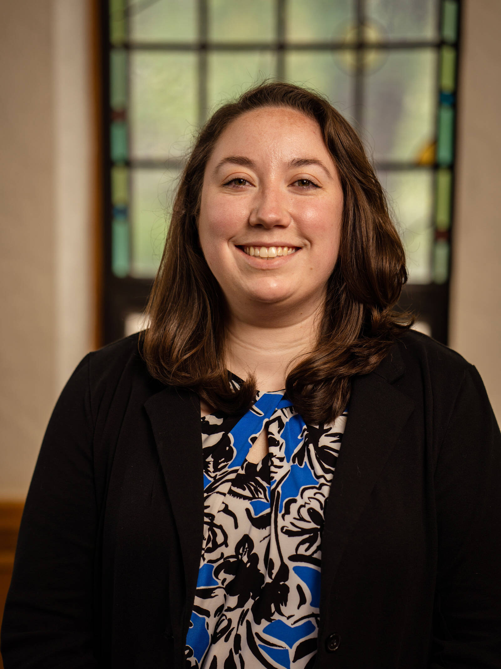 A woman with long brown hair wearing a black blazer and a blue, black, and white patterned blouse smiles while standing indoors in front of a stained glass window.