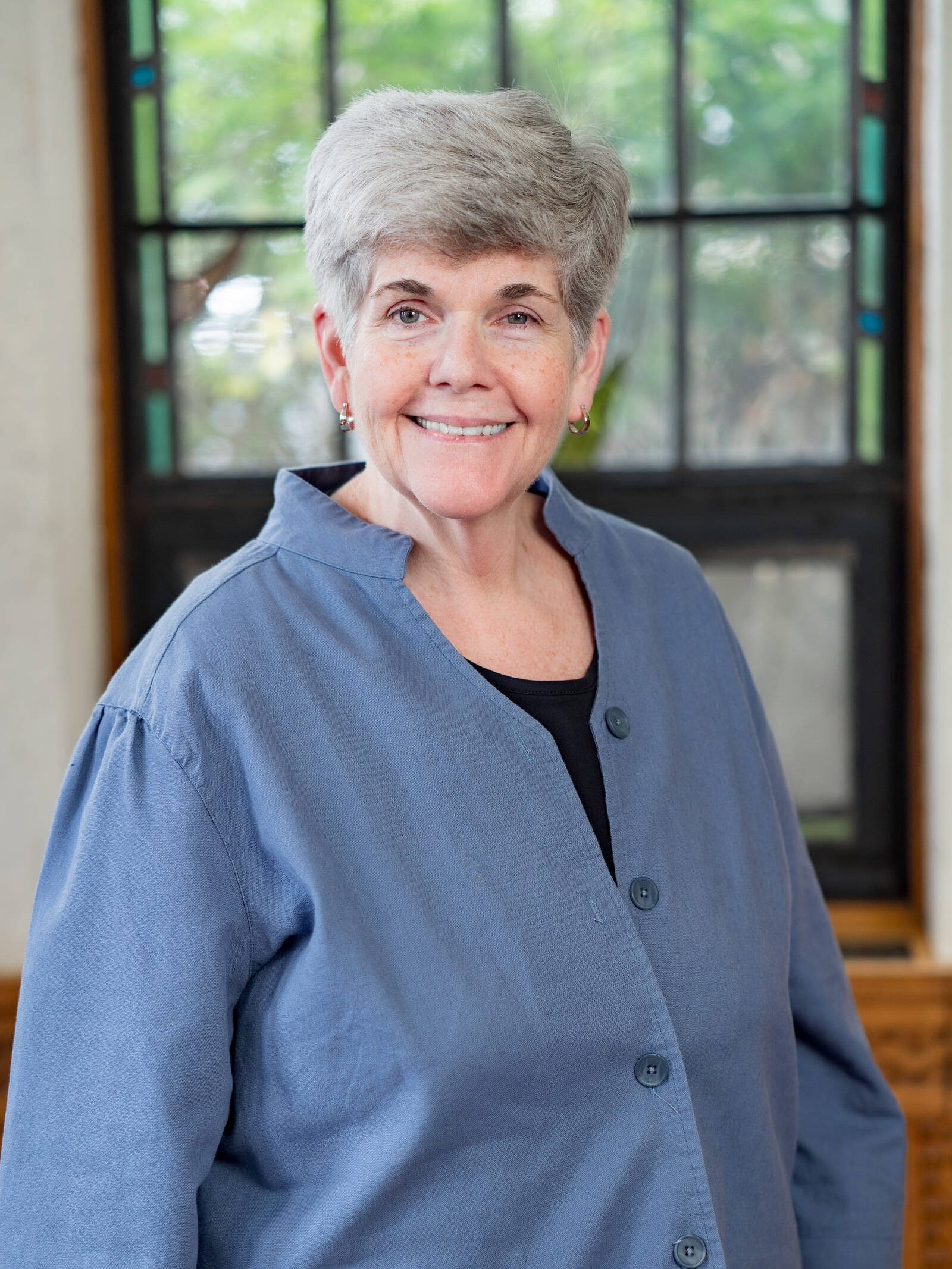 A smiling older woman with short gray hair wears a blue button-up shirt over a black top, standing indoors in front of a large window with greenery visible outside.