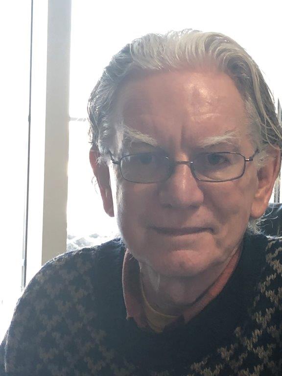 An older man with glasses and gray hair is smiling slightly at the camera. He is wearing a patterned sweater and a collared shirt. The background is softly lit by daylight from a window.