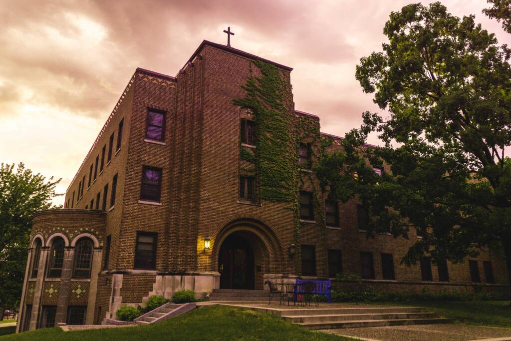 A large, historic brick building with a cross on top, partially covered in ivy, stands under a cloudy sky. The entrance features an arched doorway and round corner structure. Lush trees and a grassy slope surround the building.