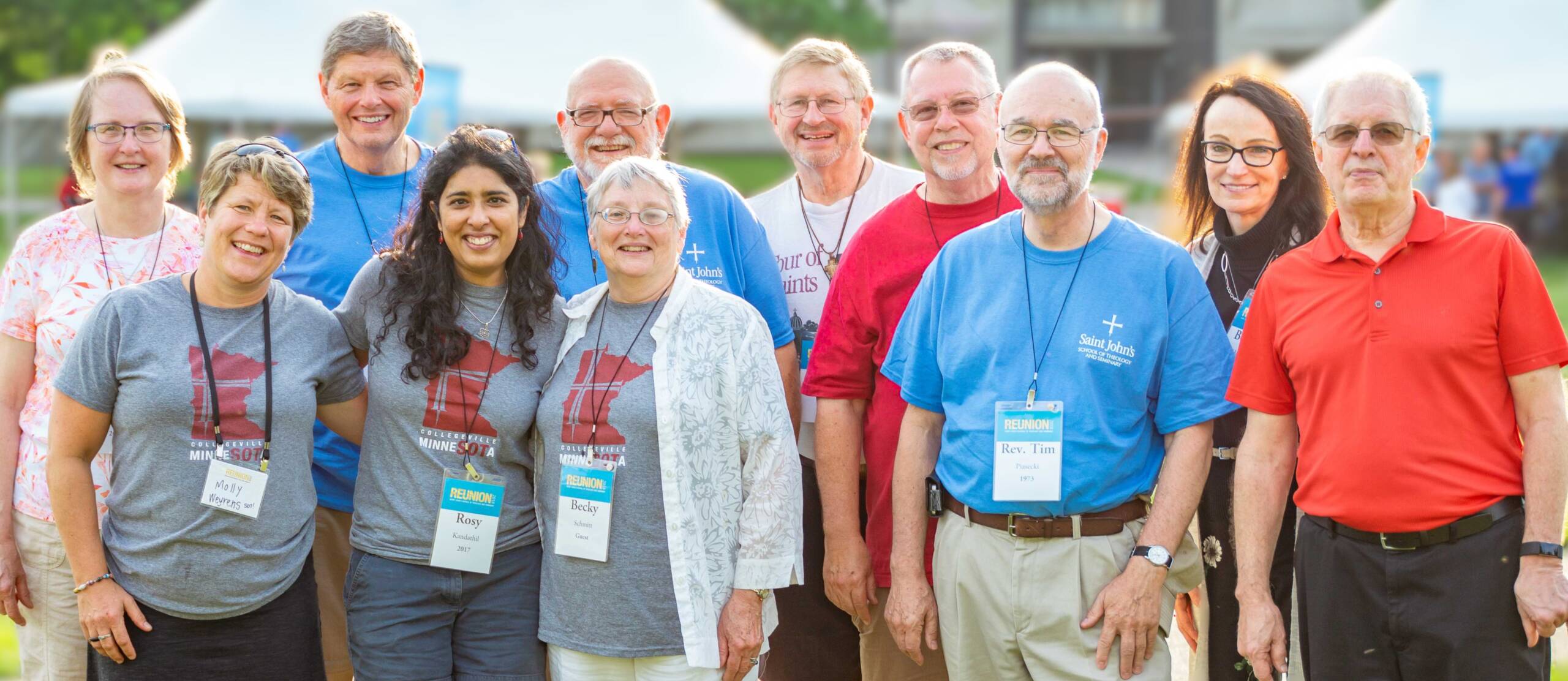 A group of people smiling and posing together outdoors, wearing name tags and colorful shirts. A white tent is visible in the background.