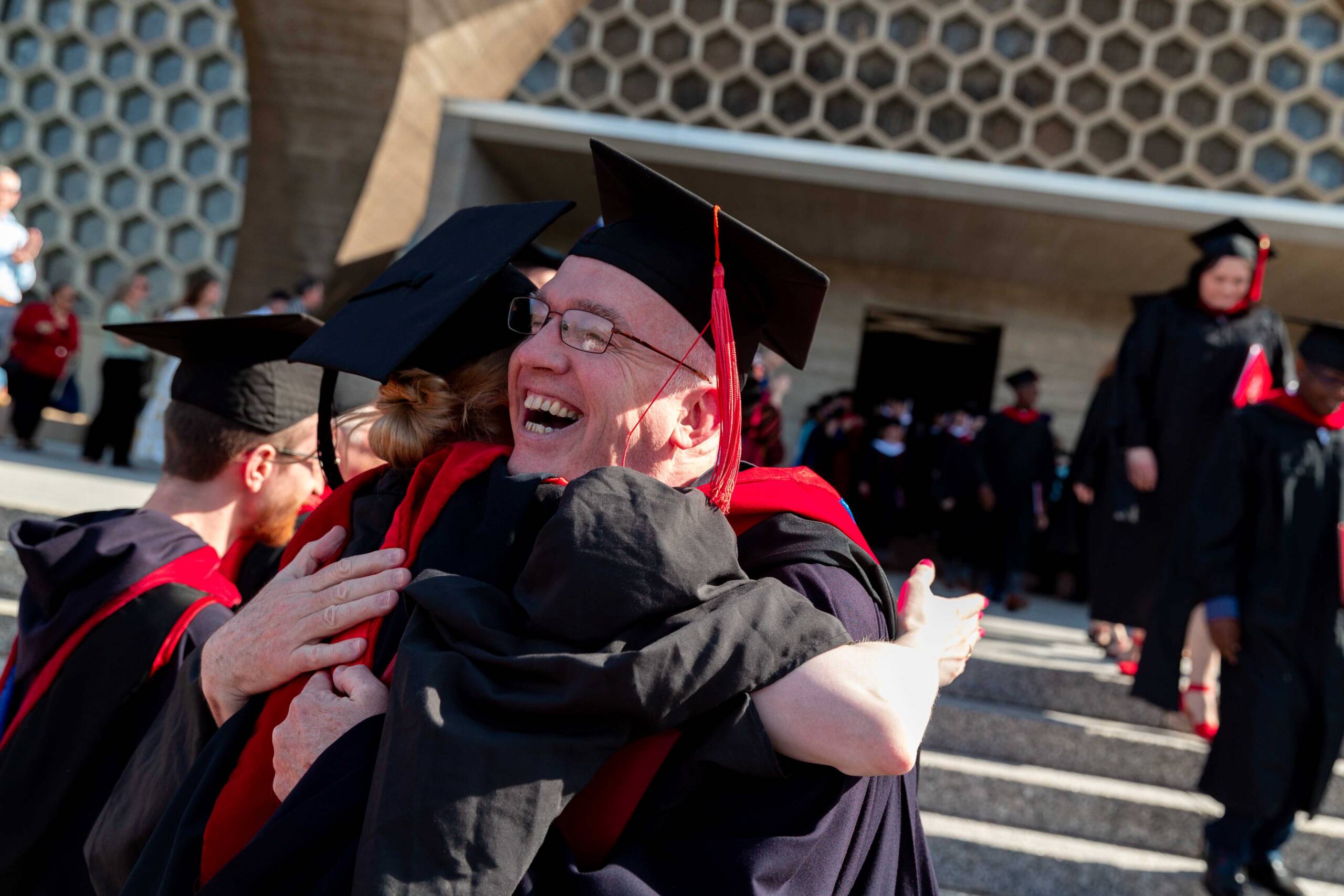 A group of graduates in caps and gowns celebrate outside a building with honeycomb-patterned architecture. They are smiling and hugging each other, conveying joy and excitement.