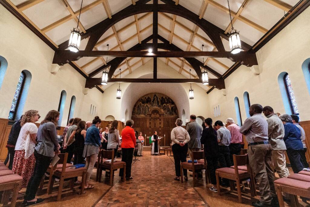 People gathered in a chapel with high ceilings and arched windows, listening to a speaker in front of an altar. The room has wooden beams and hanging lights, creating a serene and reverent atmosphere.
