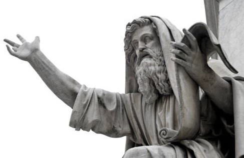 A stone statue of a bearded man wearing a flowing robe and head covering. He has one arm outstretched and the other holding a scroll. The statue is set against a light, plain background.