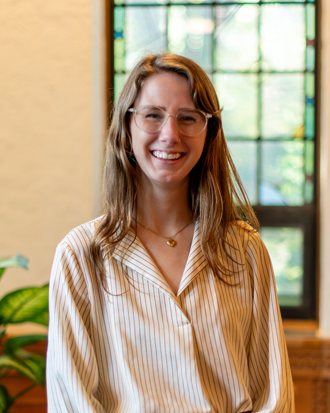 A woman with long brown hair and glasses, wearing a white blouse with thin stripes, smiles while standing indoors in front of a stained glass window. A green plant is visible to her left.