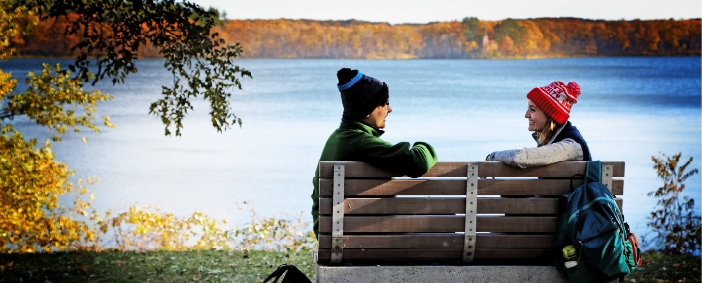 A man and a woman sit on a bench facing each other, wearing winter hats and jackets. They are near a lake, with autumn trees in the background. A backpack rests beside the woman. The scene captures a crisp, clear day.