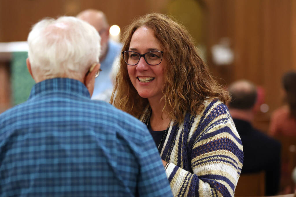 A woman with curly hair and glasses, wearing a striped sweater, smiles while talking to an elderly man wearing a blue plaid shirt. They are indoors with blurred people and a wooden wall in the background.