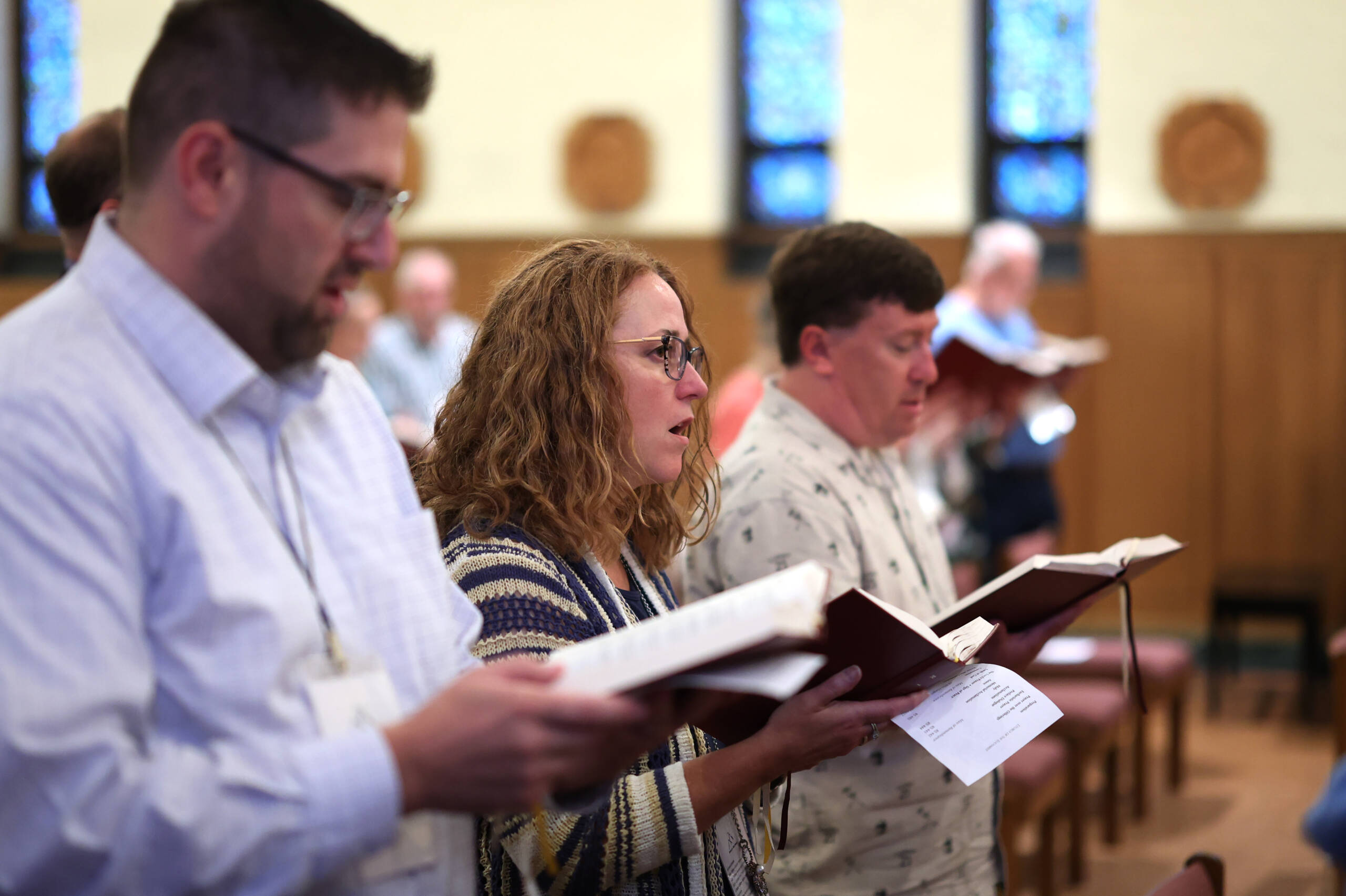 A group of people are standing in a church, holding hymnals and singing. The room has wooden pews and stained glass windows in the background. The focus is on a woman with glasses in the center.