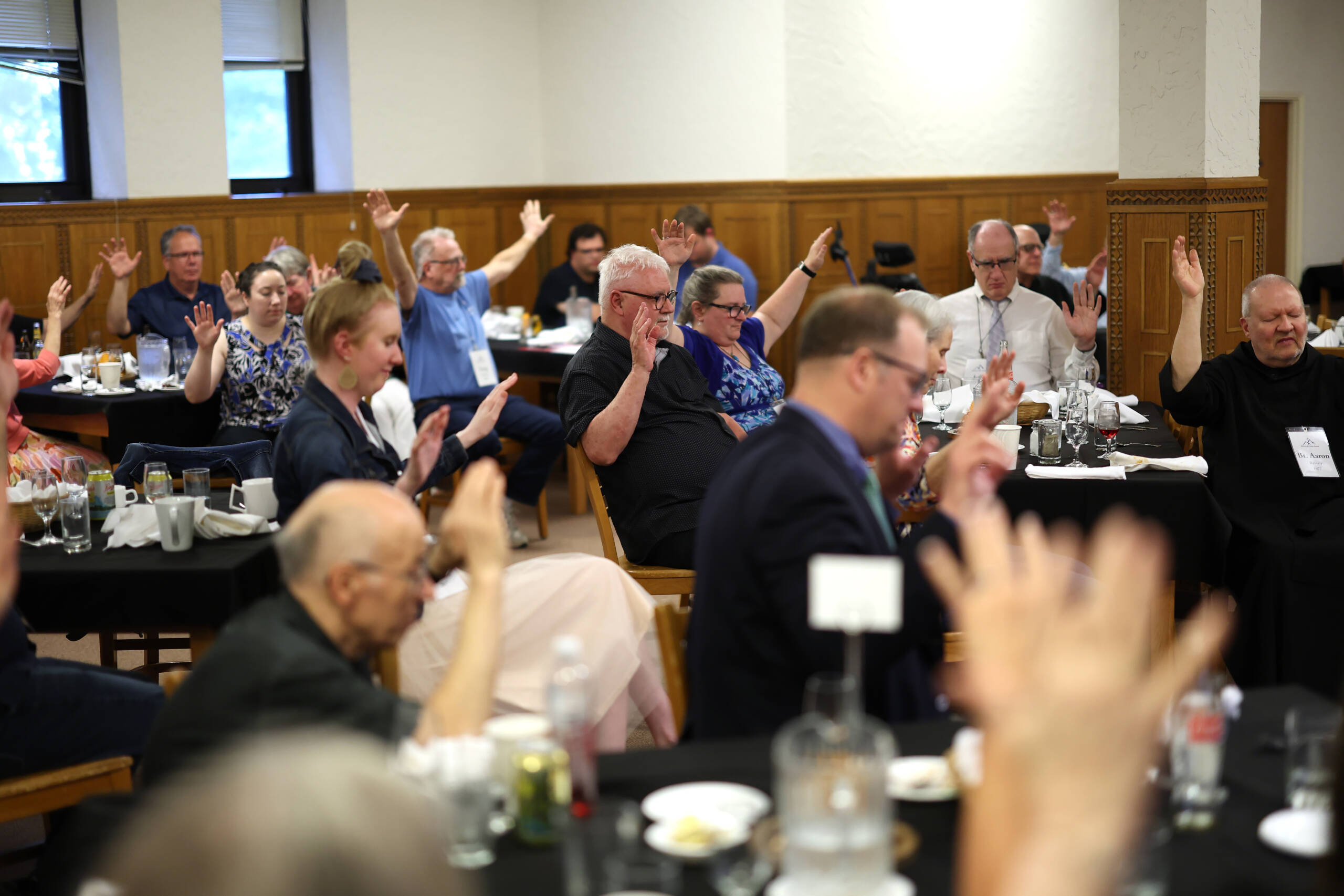 A group of people seated at tables in a conference room, raising their hands. The room has wood paneling and windows. There are plates, glasses, and bottles on the tables, and attendees are dressed in formal and casual attire.