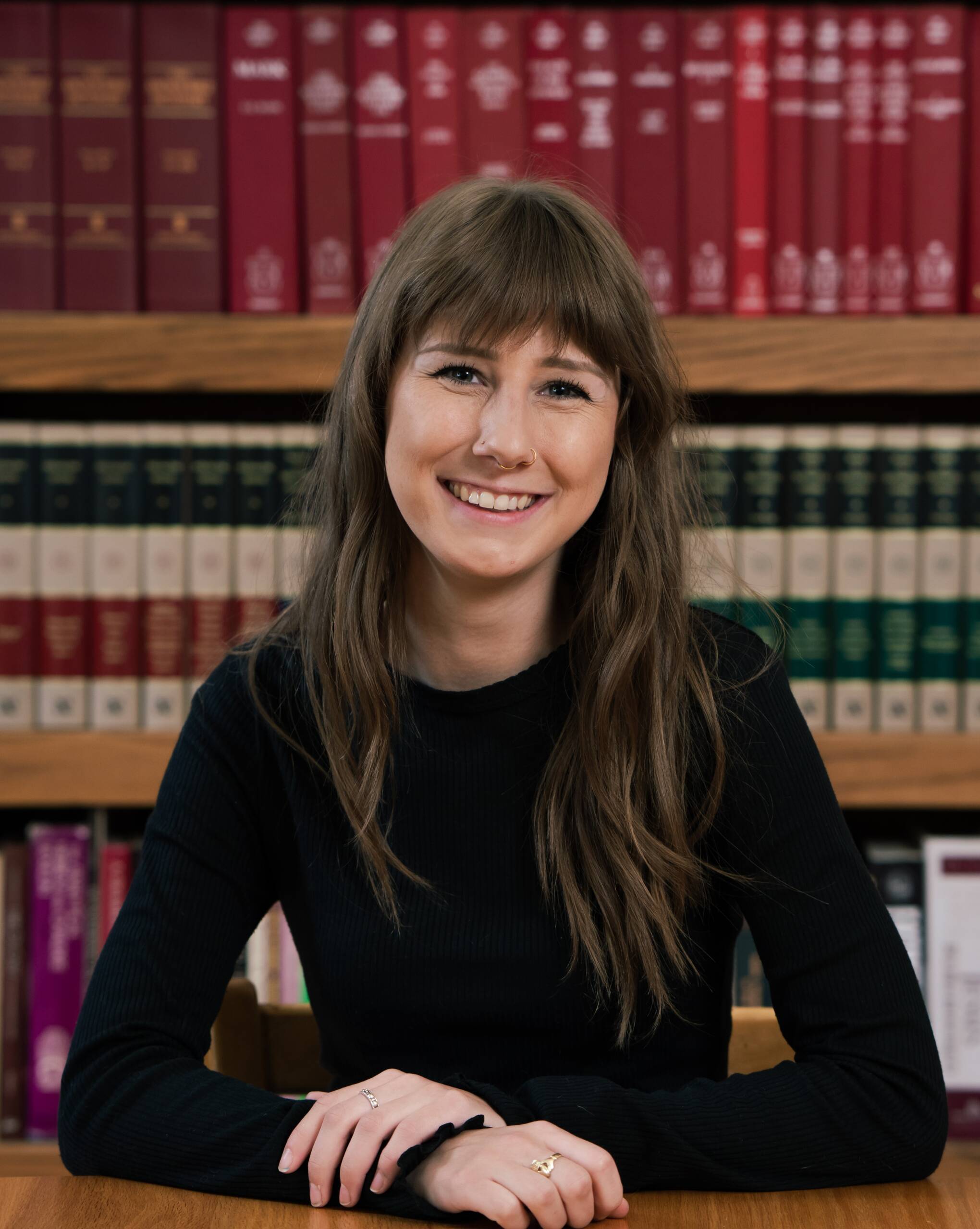 A young woman with long brown hair and bangs, wearing a black long-sleeve top, sits at a table and smiles. Behind her are shelves filled with books, including red and brown volumes.