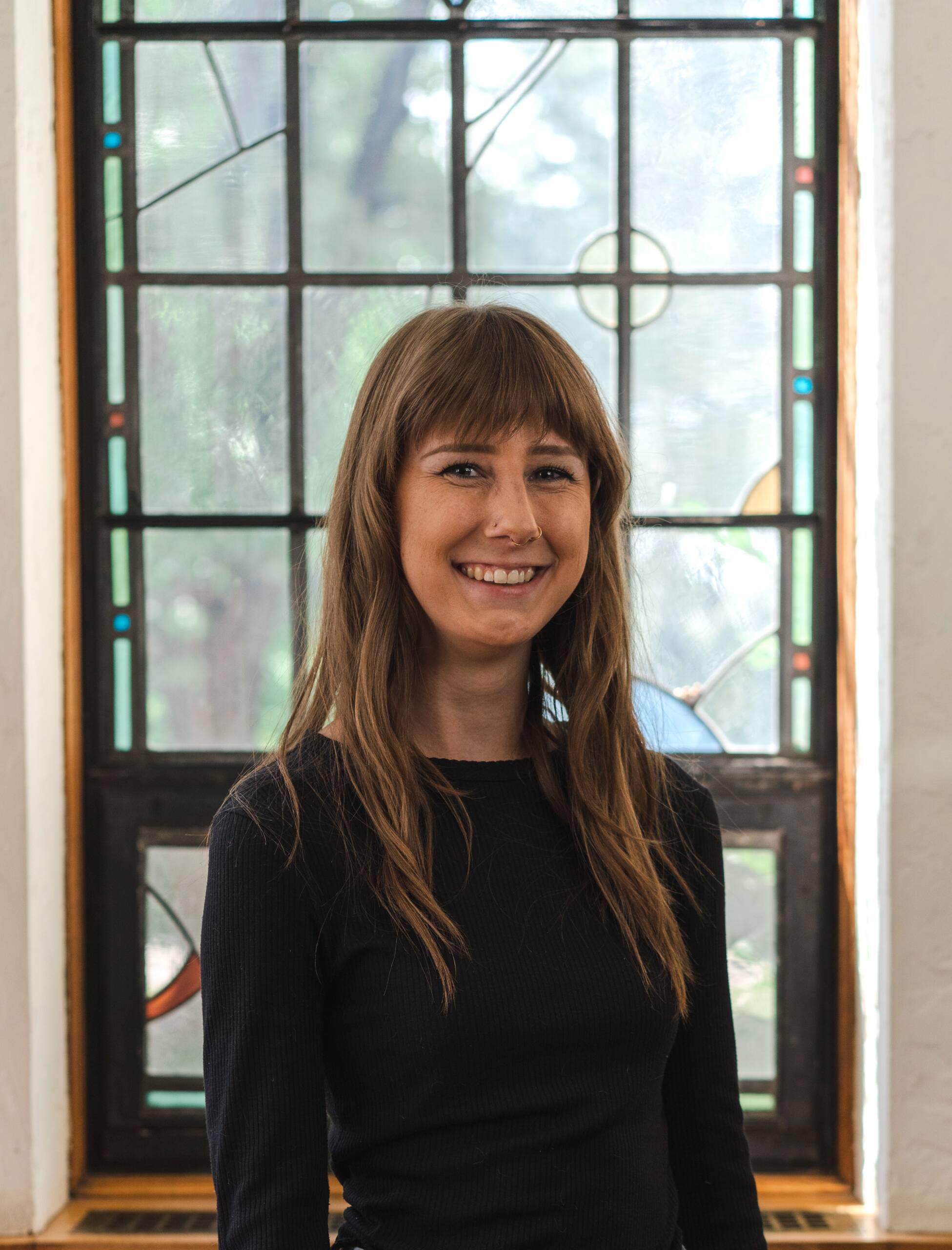 A woman with long brown hair and bangs, wearing a black long-sleeve shirt, smiles while standing in front of a stained glass window with geometric patterns.