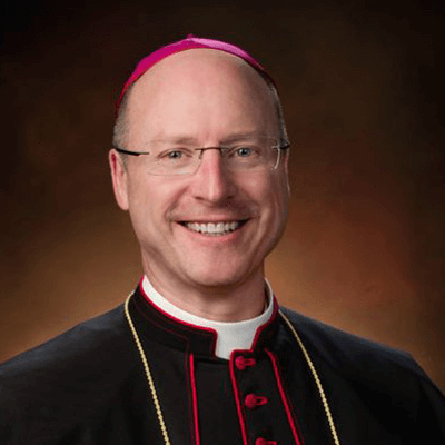 A smiling man wearing glasses, a red skullcap, and clerical robes with red piping and buttons, posing against a warm brown background.