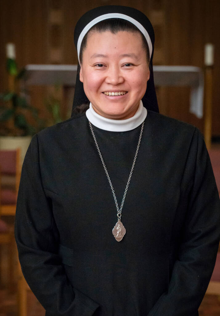 A smiling woman wearing a black and white nun’s habit and a silver pendant stands indoors, with blurred chairs and plants in the background.