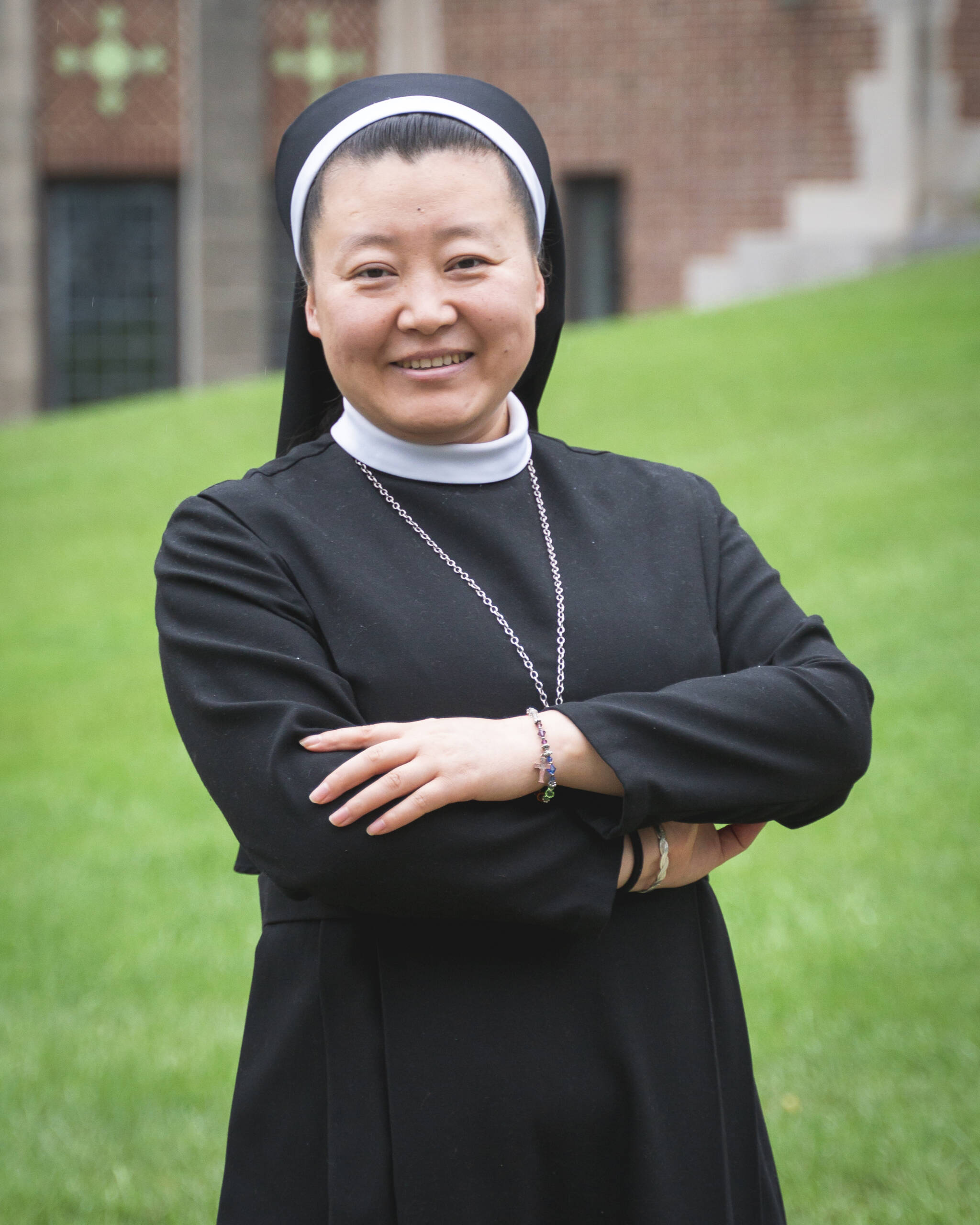 A smiling woman in a black nun’s habit and white coif stands outside on grass with arms crossed, in front of a brick building with green crosses visible in the background.
