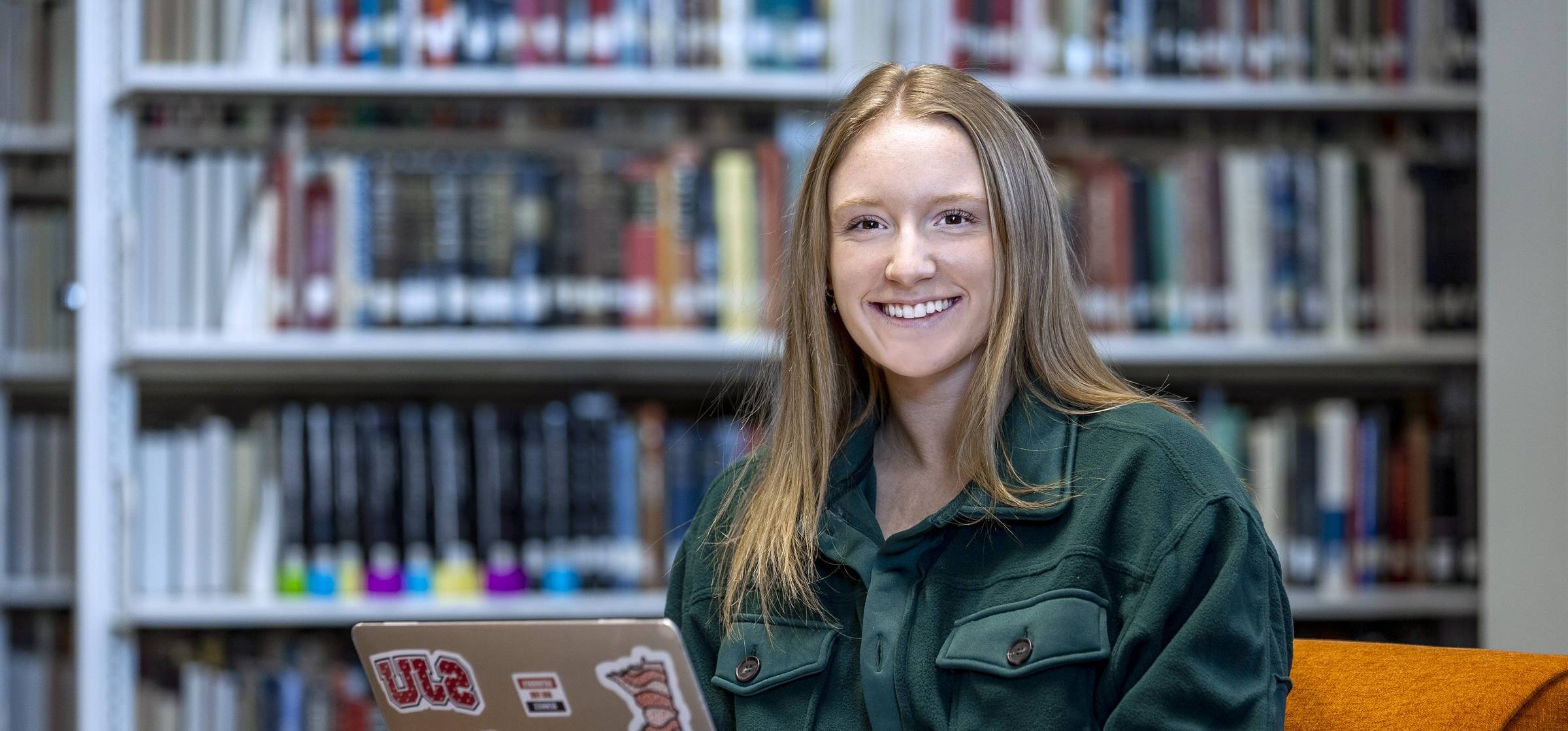 A young woman with long hair sits on an orange chair in a library, smiling while using a laptop covered in stickers. Bookshelves are visible in the background.