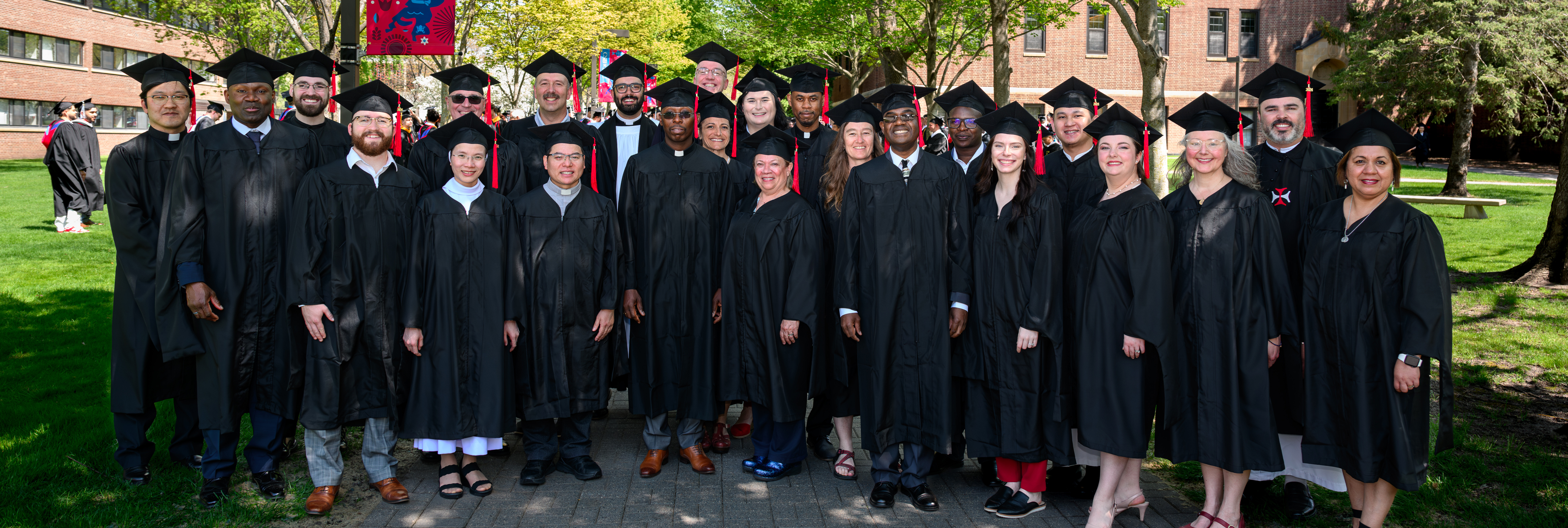 A group of graduates in black caps and gowns stand together outdoors, smiling at the camera, with trees and a brick building in the background.