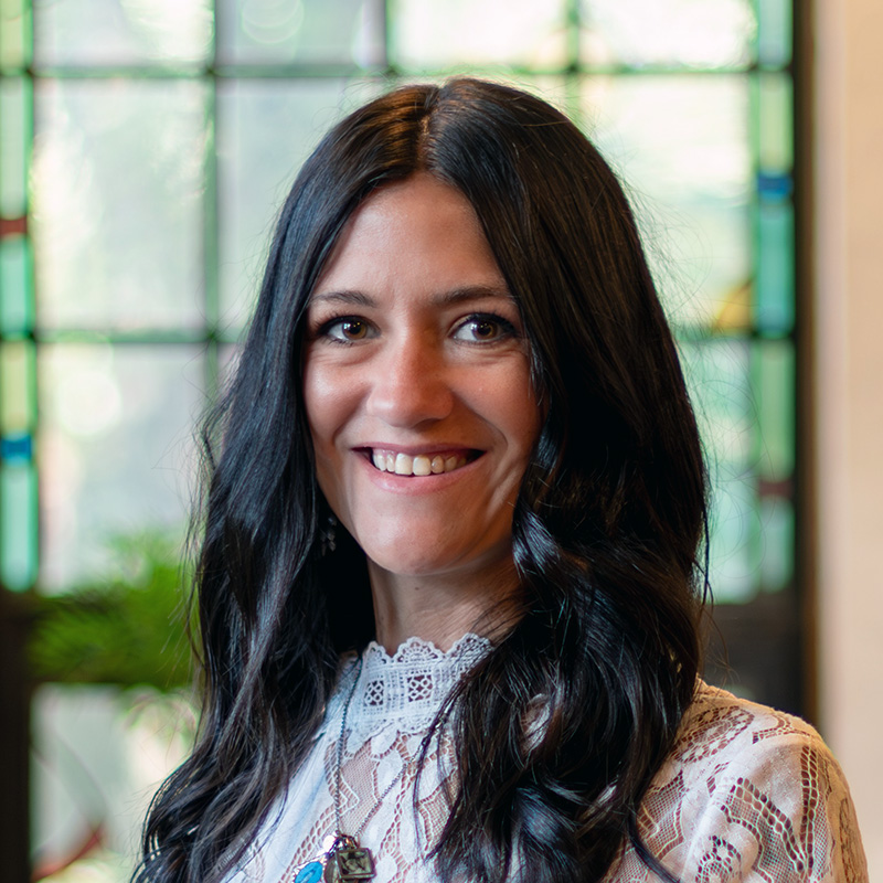 A woman with long, wavy dark hair smiles at the camera. She is wearing a lace top and stands in front of a window with colorful stained glass panels.