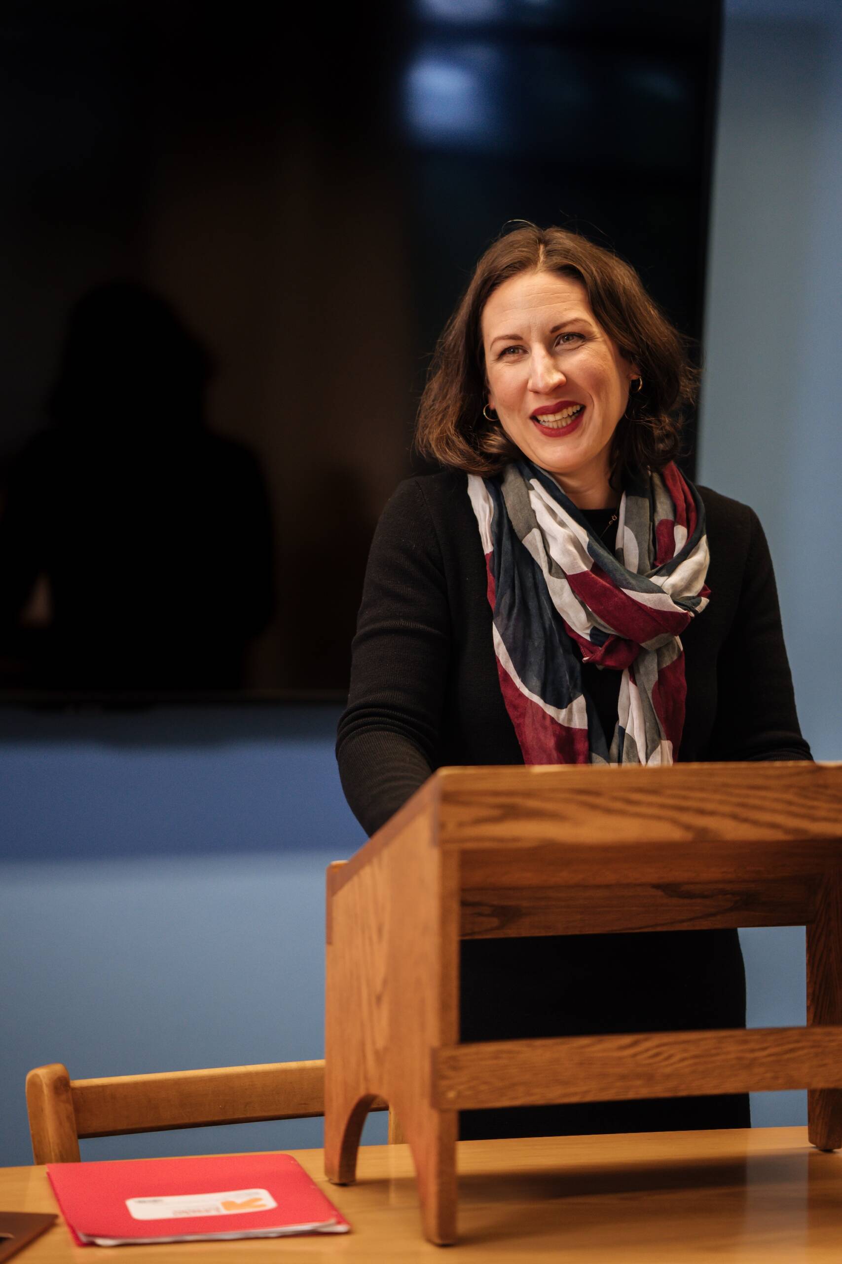 A woman with shoulder-length brown hair, wearing a black top and a red, white, and blue scarf, smiles while standing at a wooden podium in a classroom or conference room. A red folder is on a nearby table.