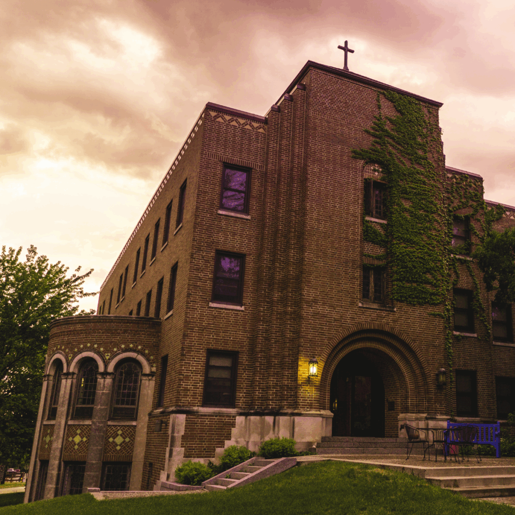 A brick building with arched windows, ivy climbing the facade, and a cross on the roof stands under a cloudy, purple-toned sky. Lit windows and a glowing porch light suggest it is evening.