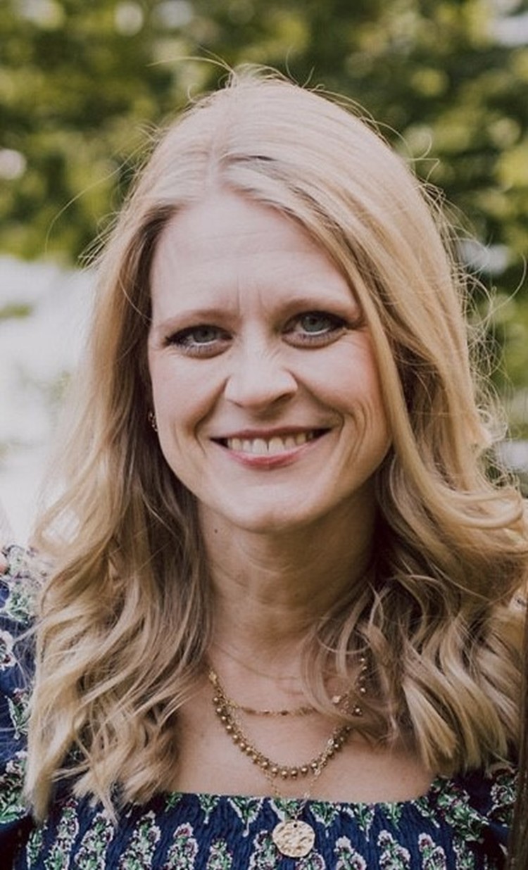 A woman with long blonde hair and blue eyes smiles outdoors. She is wearing a dark floral top and gold layered necklaces. Green trees and sunlight form a blurred background.