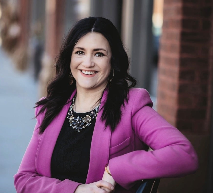 A woman with long dark hair, wearing a bright pink blazer and a statement necklace, smiles while leaning on a chair outdoors near a brick wall.