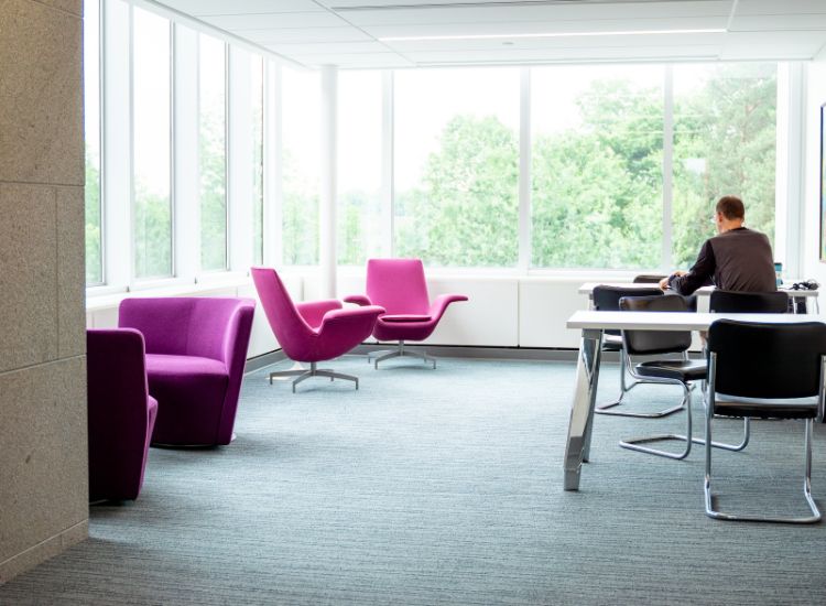 A bright, modern office space with large windows, purple lounge chairs, and a person sitting alone at a table working. Green trees are visible outside.