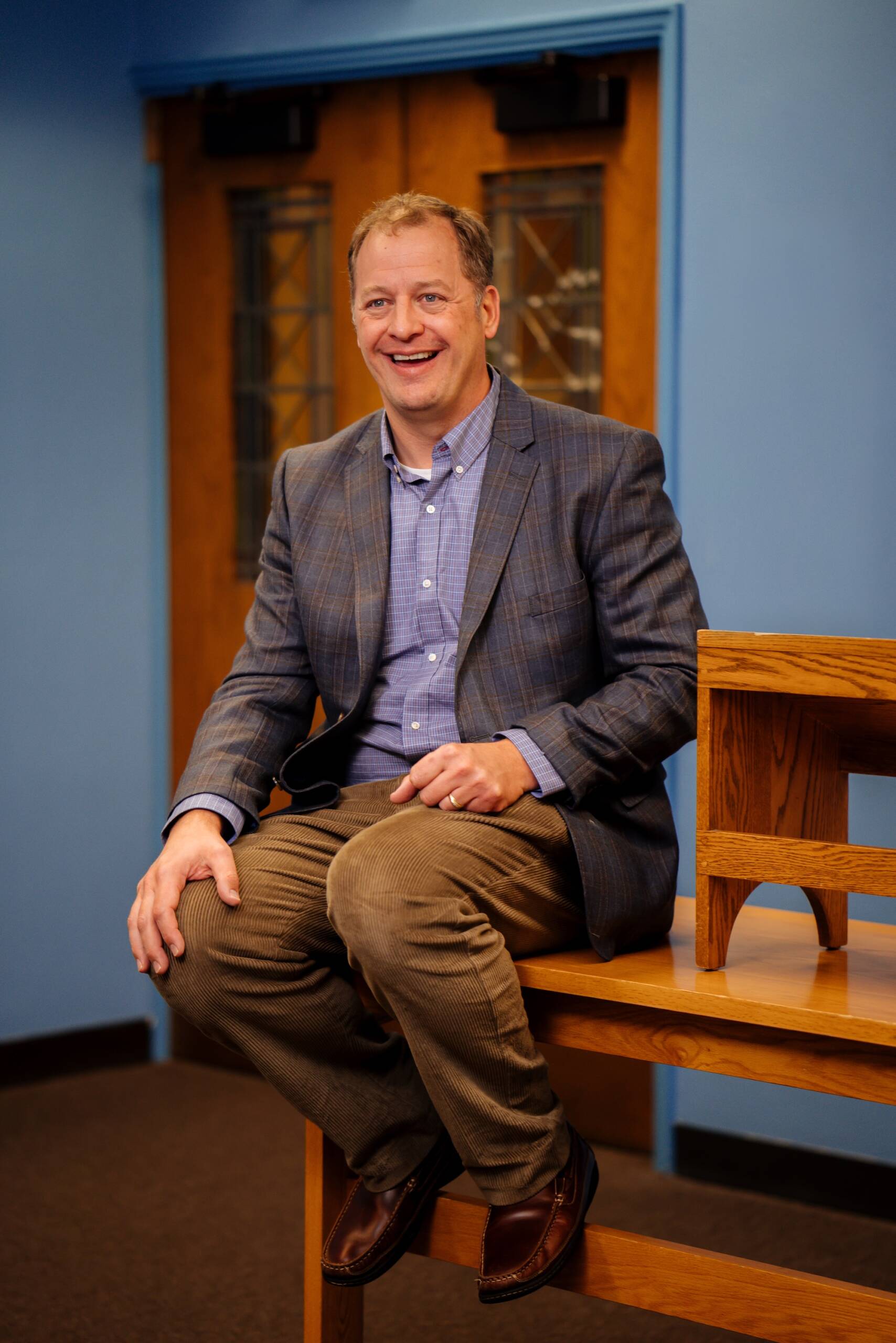 A smiling man in a plaid blazer, blue shirt, and brown pants sits on the edge of a wooden table in a room with blue walls and wooden doors in the background.