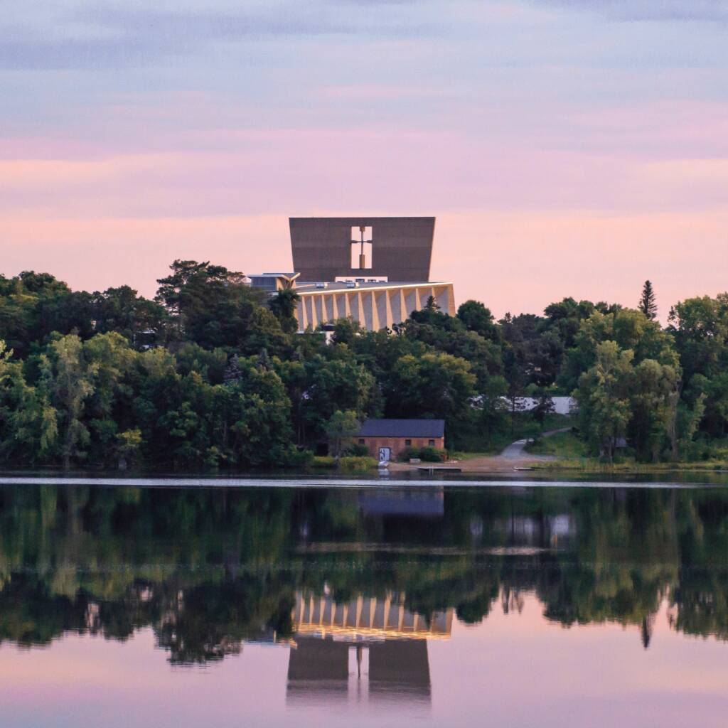 A modern building with a cross on its facade rises above a line of trees near a calm lake, reflecting the building and greenery in the water under a pastel sunset sky.